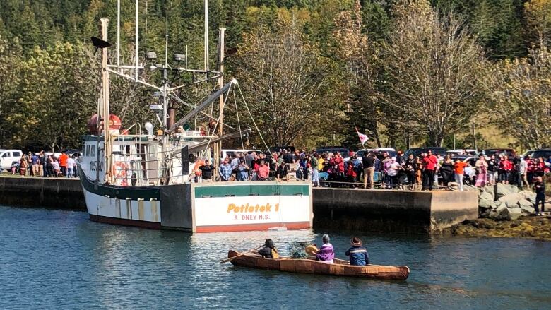 People in a canoe paddle past a large fishing boat tied up in a canal lined with people.