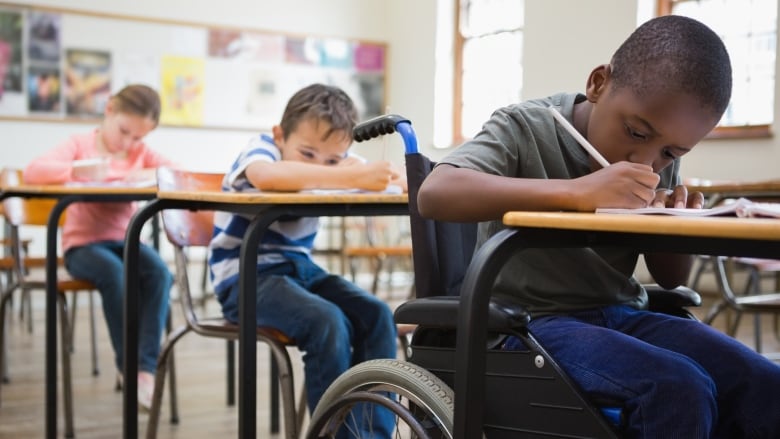 Young students are seen writing at desks in classroom, with the student at right seated in a wheelchair.