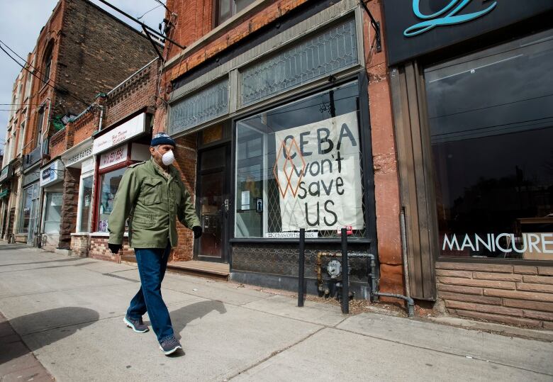 A person wearing a mask walks by storefronts, including one that displays a sign that says, 'CEBA won't save us.'