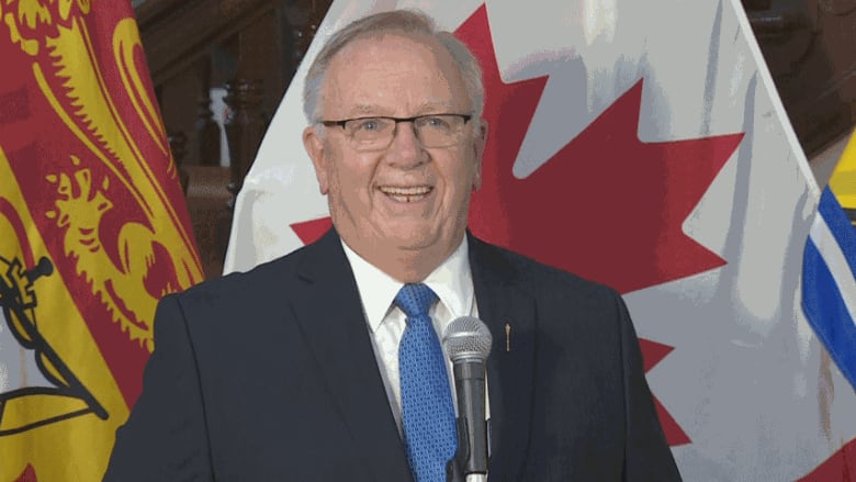 A smiling man in a black suit and blue tie in front of a microphone. New Brunswick and Canada flags are in the background.