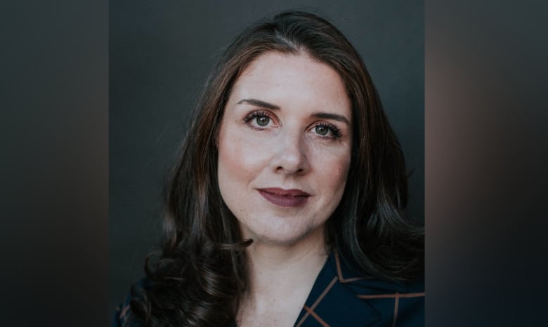 Gillian Hnatiw is wearing a black v-neck shirt and is sitting in front of a dark background. She has long dark brown hair.