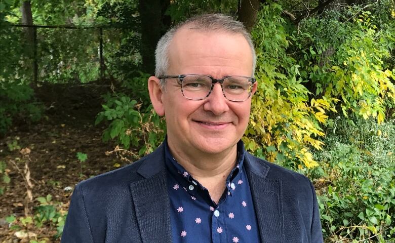 A man poses for a photo outside in front of a leafy backdrop.