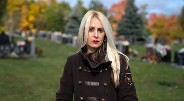 A middle-aged blond woman in a park wears fall clothing. She is looking at the camera, visible from the waist up, with hands in coat pockets.