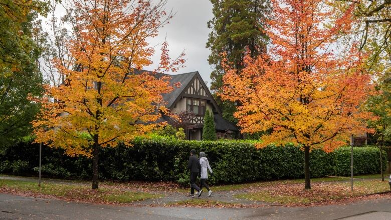 People walk past a large home with autumnal leaves falling around them.