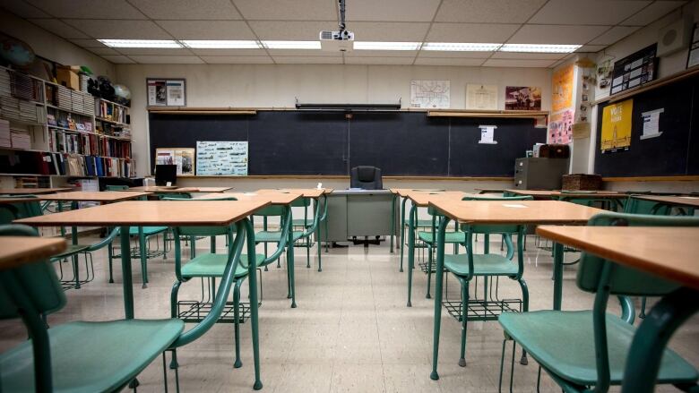 Several green desks with wooden tops sit in an empty classroom facing the board.