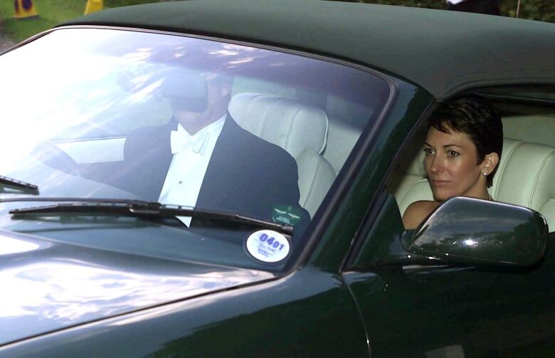 A man in a tuxedo drives a car while a woman with short black hair sits in the passenger seat.