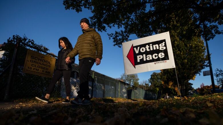 Two people holding hands walk past a sign that reads 'Voting Place' with an arrow pointing left.