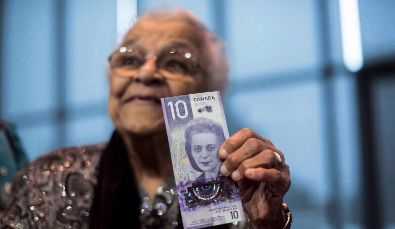 An older Black woman holds a 10 dollar bill with the face of Viola Desmond on it