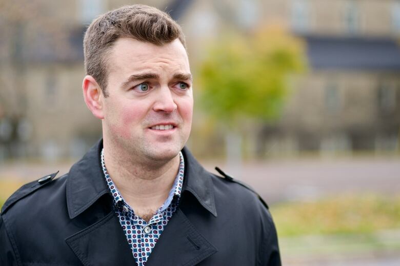 A man wearing a checkered shirt with a black coat overtop. The outdoor background is blurred.