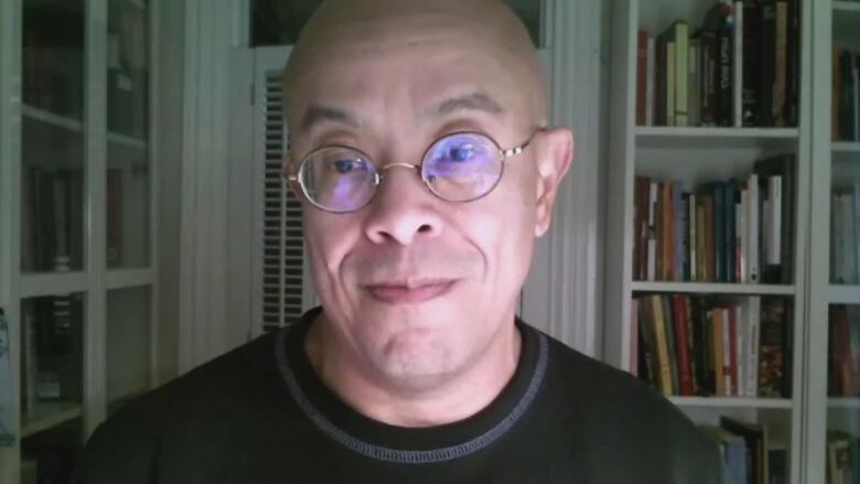 A bald man wearing a black shirt and glasses sitting in front of a bookshelf