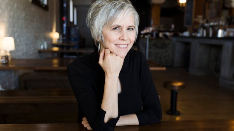 Shauna Powers sits in a restaurant with low lighting, dark wooden tables and white stone walls. She has white hair and is wearing a black cardigan.