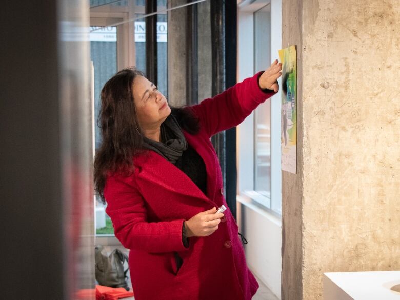 A woman with dark hair in a red coast puts up a small poster beside a plywood facade.