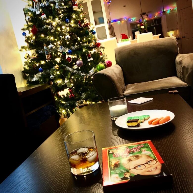 A plate of Christmas cookies sits on a coffee table alongside a glass of milk. A decorated Christmas tree is seen in the background.