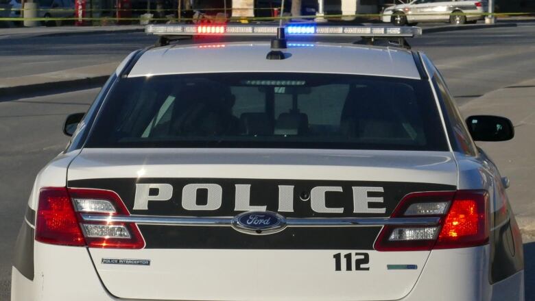 A Winnipeg police car, from the back. It is mostly white with black trim. The word, Police, is written across the trunk. The red and blue lights on the roof are flashing.