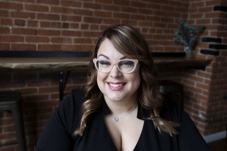 Sam Maciag sits in front of a red brick wall. She has long brown hair and beige glasses. She is wearing a black cardigan.