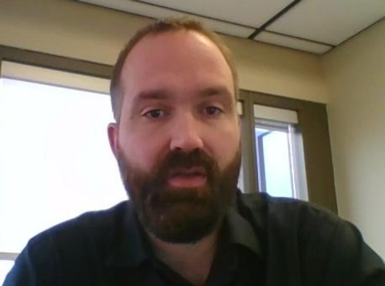 A portrait of a man with dark hair and a beard, wearing a black collared shirt.