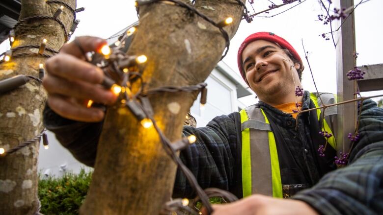 A man smiles as he wraps Christmas lights around an exterior branch.