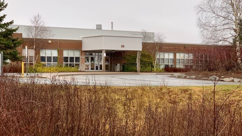 The main entrance of a single story school is seen. The walls are made of red-brown brick and the front doors are under a metal awning.