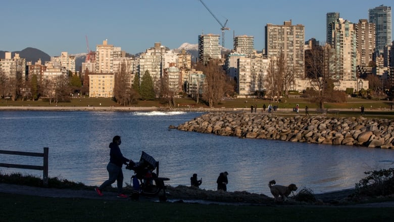 A person pushing a stroller is silhouetted in front of the ocean and a city skyline on a sunny day.