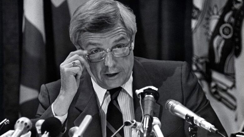 A politician lowers his glasses as he sits behind several microphones, in a black-and-white photo of former Alberta premier Peter Lougheed from 1984.