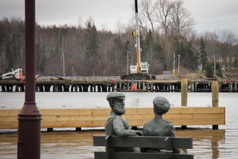 A statue of two people sitting on a park bench seen in foreground, with water and a large dock in background.