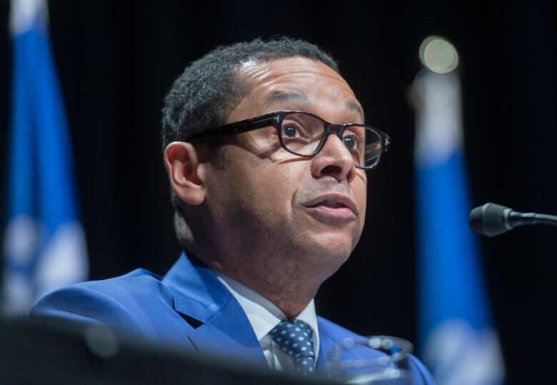 A man sitting in front of Quebec flags speaks into a microphone. 