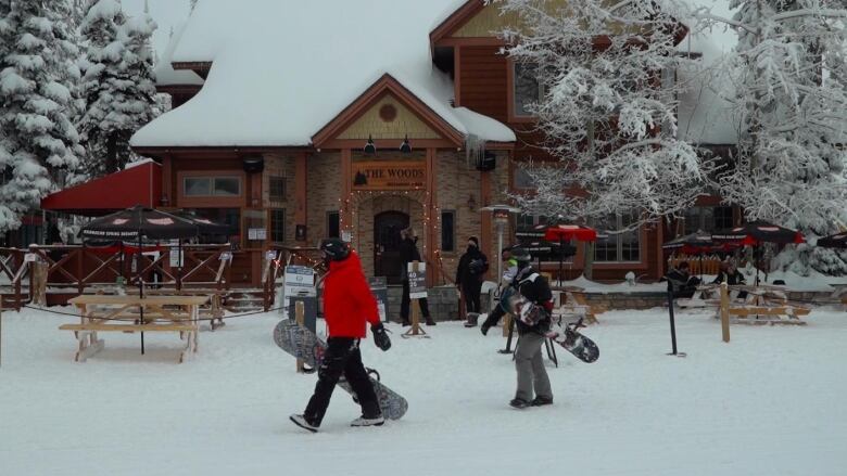 Two snowboarders walk past a lodge in the snow.