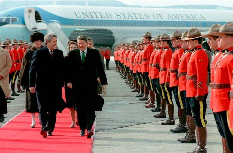 Two men walk down a red carpet past rows of RCMP officers in red serges.