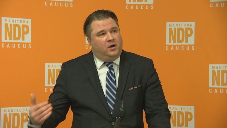 A man in a suit stands in front of an orange backdrop with the Manitoba NDP caucus logo on it.