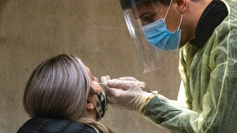 A woman tilts her head back as a male health-care worker wearing full protective gear inserts a long swab in her nostril. 