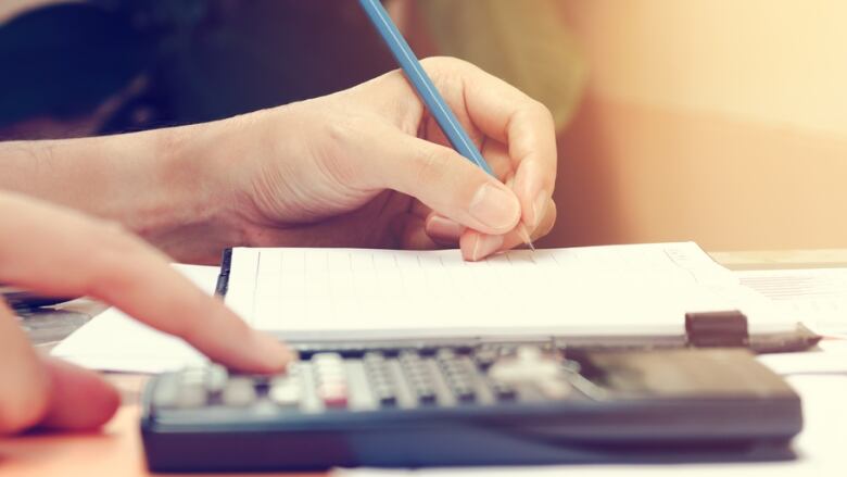 Two hands are seen in a closeup image, one holding a blue pencil above a notepad and the other pushing keys on a calculator.