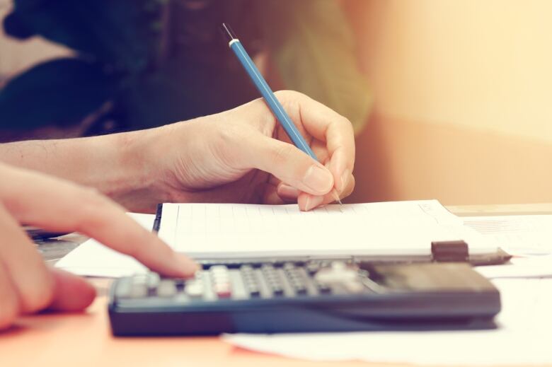 Two hands are seen in a closeup image, one holding a blue pencil above a notepad and the other pushing keys on a calculator.