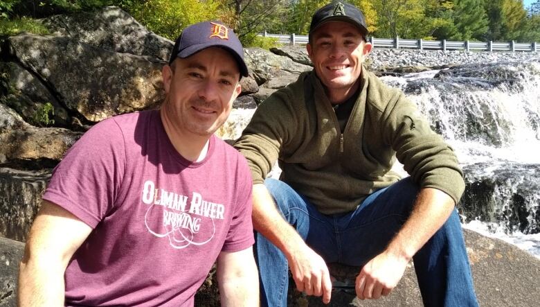 Two men sit on rocks in front of a waterfall. 