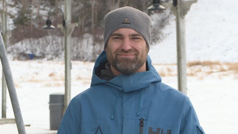 A man wearing a blue winter jacket and a grey winter hat standing in front of a ski hill covered in snow. 