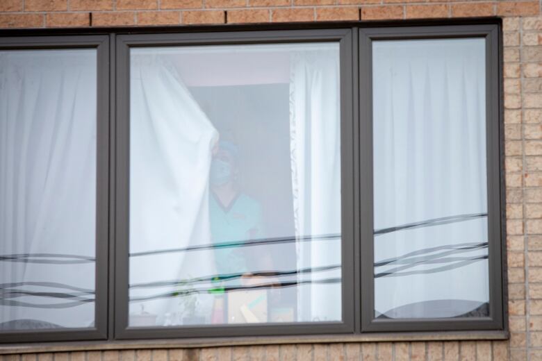 A long-term care staff member wearing personal protective equipment is seen through a window pulling curtains closed. 