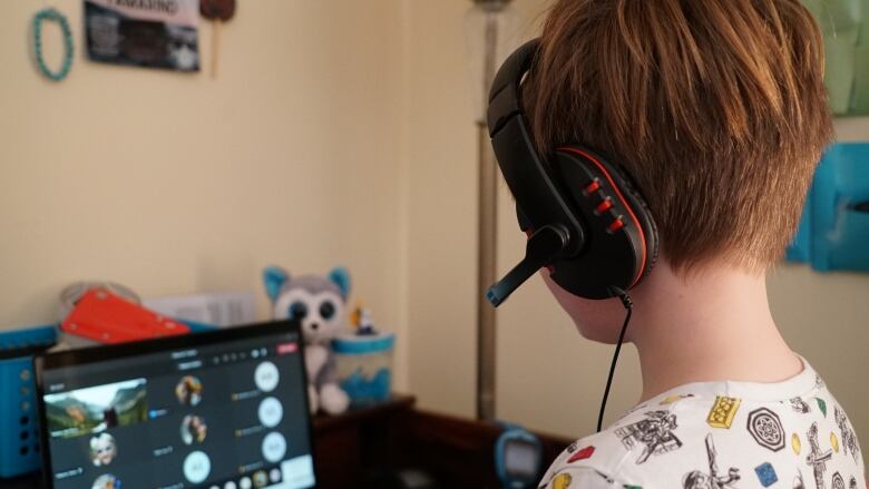 A child, shown from behind wearing a microphone and headphones computer headset, looks down at a laptop computer logged into an online classroom.