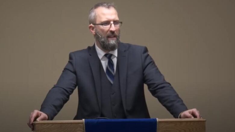 a man in a grey suit and beard speaks at a podium 