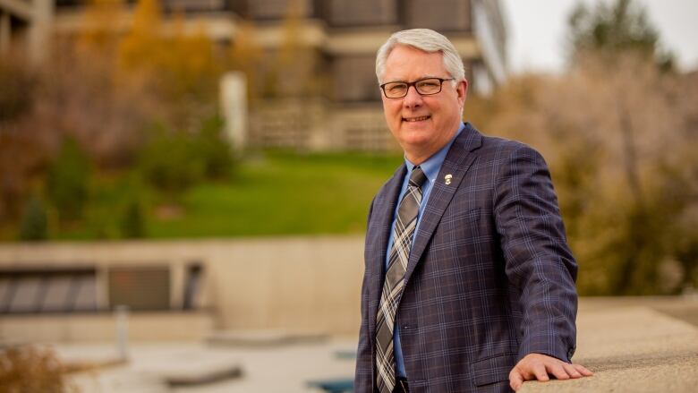 Mike Mahon, a grey-haired man with classes in a business suit, stands in front of a college building surrounded by trees.