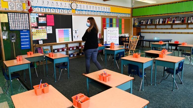 A female teacher walks past empty desks in a classroom. 