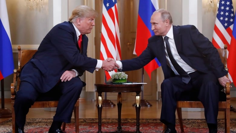 Two men shake hands across a table, with flags in the background.