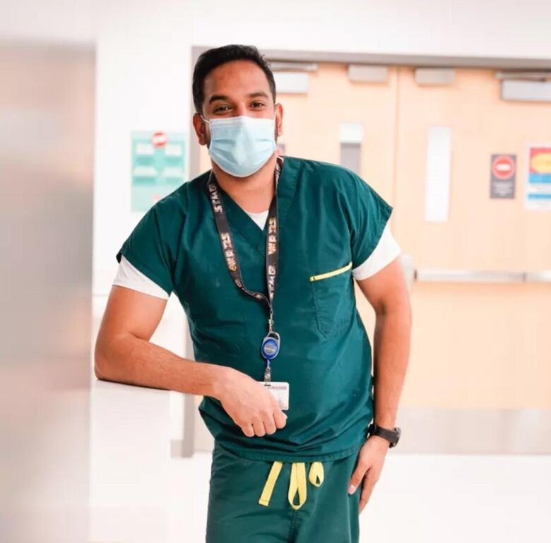 A man wearing green scrubs and a mask leans against a wall. 