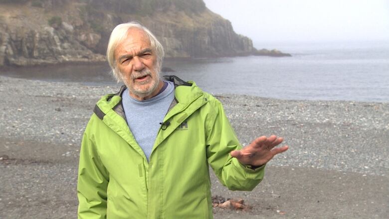 A white-haired bearded man stands on a rocky beach.