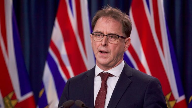 A white man speaks at a podium, with two B.C. flags behind him.