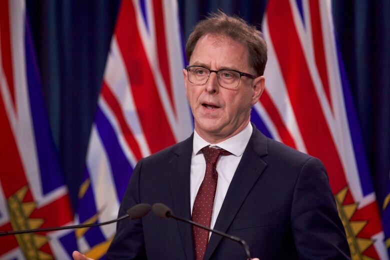 A white man speaks at a podium, with two B.C. flags behind him.