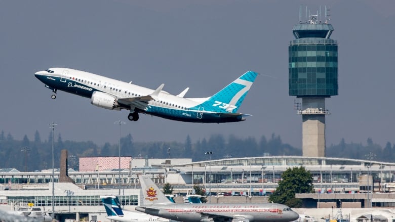 A Boeing 737 Max 8 airplane, with a white and dark blue body and turquoise tail, takes off from an airport, lifting off in front of an air traffic control tower. 