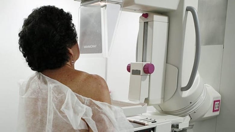 A woman with short black hair wearing a hospital gown is shown from behind as she undergoes a mammogram.