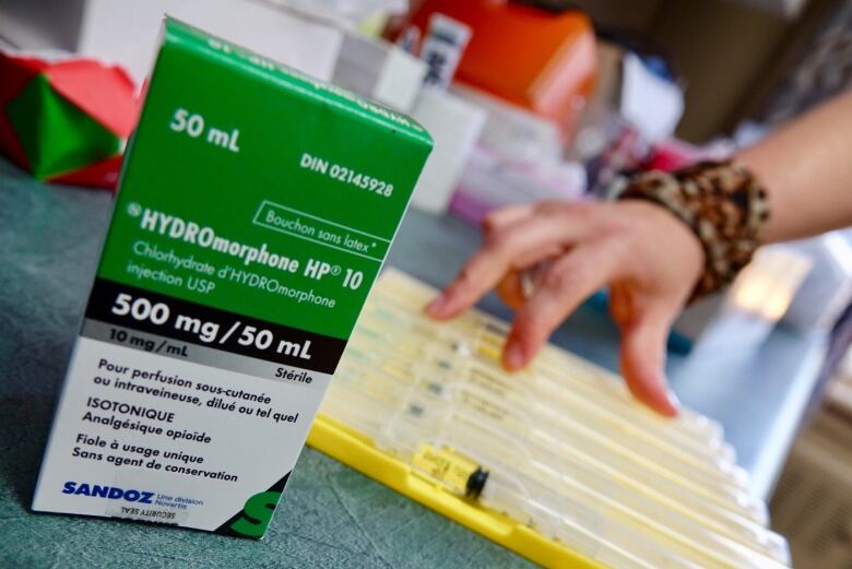 A green box of medication is pictured. A person's hand is visible in the background with  a tray of medical supplies.