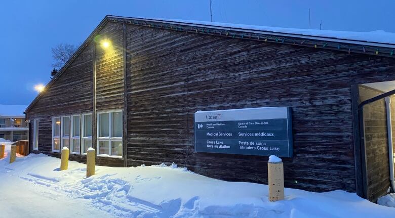 A timber A-frame building is shown surrounded by snow. There's a light on in the front to illuminate the building as darkness falls.