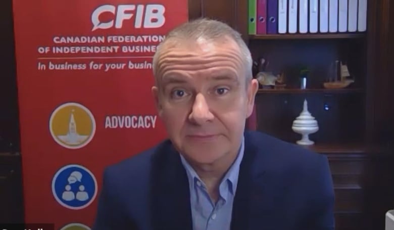 A man in a collared shirt and jacket sits in front of a poster that reads 