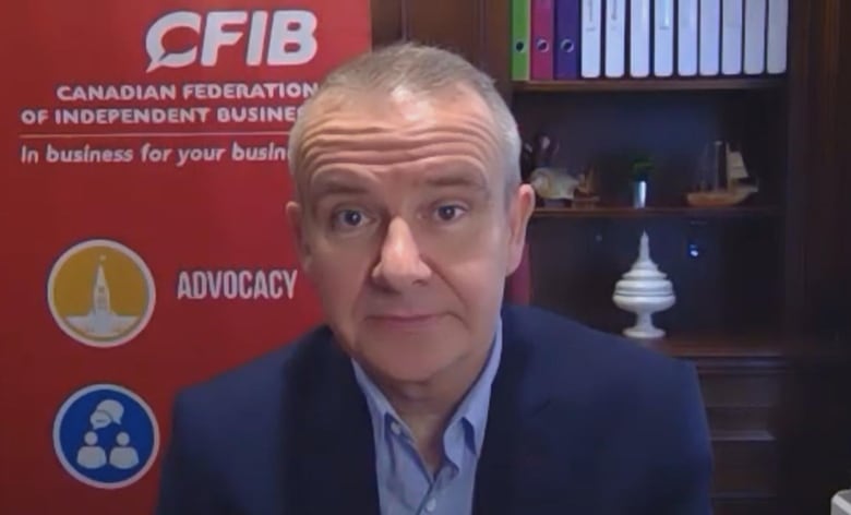 A man in a collared shirt and jacket sits in front of a poster that reads 